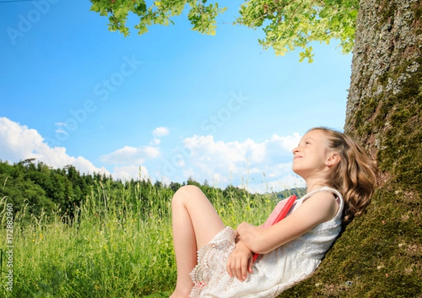 Obraz young girl sit next to a tree and smile into the summer sun. she plays with electronic devices, red apple, flowers. sky is blue and the grass is green.