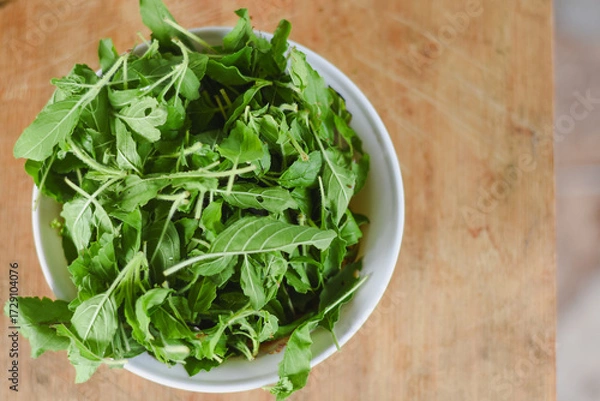 Fototapeta overhead shot focusing on a white bowl filled with fresh, bright green leaves placed on a light wooden surface.