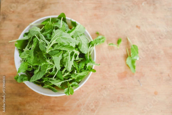 Fototapeta overhead shot focusing on a white bowl filled with fresh, bright green leaves placed on a light wooden surface.