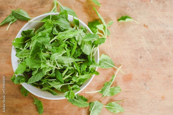 Fototapeta overhead shot focusing on a white bowl filled with fresh, bright green leaves placed on a light wooden surface.