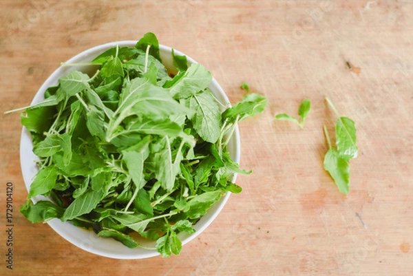 Fototapeta overhead shot focusing on a white bowl filled with fresh, bright green leaves placed on a light wooden surface.