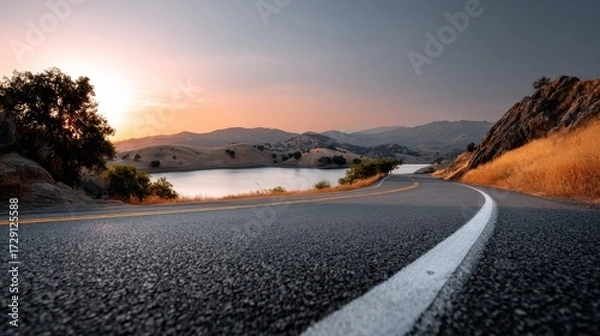 Fototapeta A winding road curves alongside a tranquil lake, framed by hills and bathed in warm sunset light, creating a serene landscape.