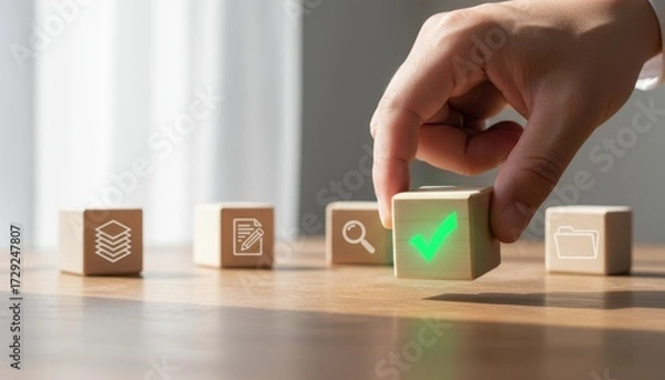 Fototapeta Complete & Correct: A Hand's Touch: A hand meticulously places a wooden cube bearing a glowing green checkmark upon a table of wooden blocks marked with the icons of a work environment.