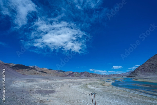 Fototapeta A vivid landscape featuring a meandering river cutting through a barren high-altitude desert, set against colorful mountains under a deep blue sky with scattered clouds. Himalayan ackground