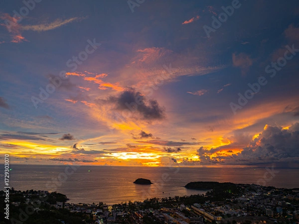 Fototapeta A breathtaking sunset paints the sky in vivid hues of orange and blue over a coastal town, with lush green hills framing the scene. A distant hilltop structure adds a serene tranquil landscape.