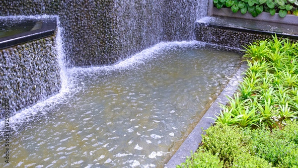 Fototapeta Waterfall curtain stone with green ornamental plant in public park garden, perspective side view with copy space