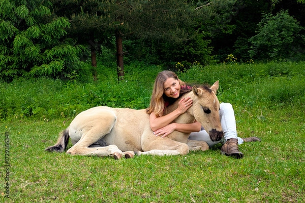 Obraz Young woman sitting on ground next to dunn color small horse foal, another animal near in background