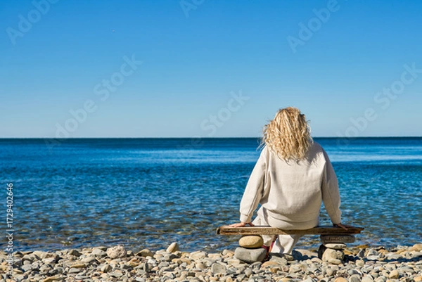 Fototapeta A rear view of a mature blonde woman relaxing on the seashore, admiring the tranquil atmosphere and enjoying moments of relaxation, copy space for the concept of outdoor recreation and sea ecosystems