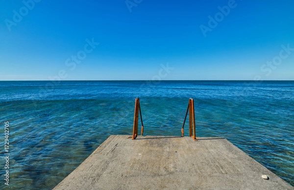 Fototapeta Pier with rusty railings for descent into the sea, clear calm weather with a view of the calm sea, background idea about marine ecosystems and human impact