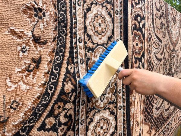 Fototapeta A close-up of a hand holding a brush, scrubbing a hanging carpet outdoors.