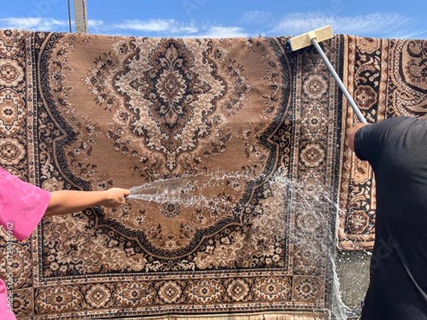 Fototapeta A traditional carpet being washed outdoors with a hose and scrub brush. A woman's hand and a man's hand are seen from opposite sides, actively cleaning the rug with water.