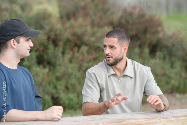 Fototapeta Two men are sitting at a picnic table, one wearing a black hat. They are talking to each other