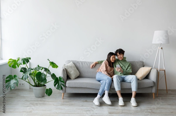 Fototapeta Happy millennial Asian couple using cellphone, texting and browsing internet, relaxing on couch indoors, copy space. Relaxed young family using mobile application, resting at home