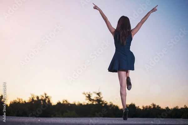 Fototapeta A young slender girl with sexy legs in heels in a miniskirt stands on a sunset background with her hands raised on one leg depicting joyful and vivid emotions.