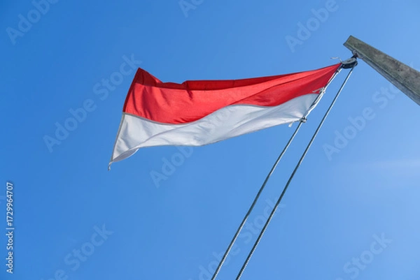 Obraz Indonesian flag waving proudly on a tall wooden pole against a clear blue sky, symbolizing independence, unity, and national pride with its red and white colors standing out brightly.