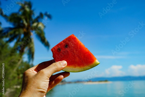 Obraz Vibrant watermelon slice held by a woman on a sunny beach with blue sky. A refreshing slice of watermelon held against a serene beach backdrop. summer bliss, vacation vibes, travel, lifestyle.
