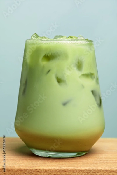 Fototapeta Iced matcha latte in transparent glass with ice on wooden surface against blue background, vertical closeup, no logo, copy space.