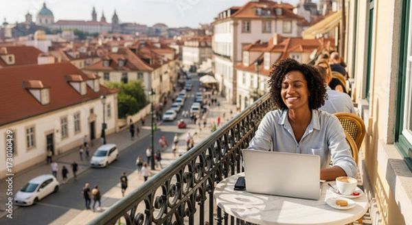 Fototapeta  A young professional (30s, diverse) is working remotely on a laptop from a scenic cafe terrace overlooking a bustling, charming European city. They are smiling, enjoying the freedom and flexibility .