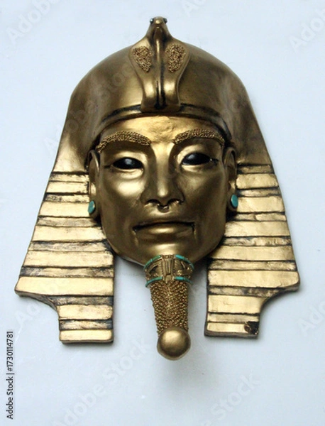 Fototapeta Ancient pharaoh mask sculpture with golden metallic finish and turquoise details. Alien-like eyes evoke mystery, blending Egyptian heritage with otherworldly symbolism.Frontal face on white background