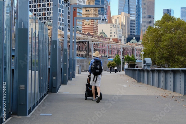 Fototapeta A father pushes a stroller across a pedestrian bridge with Melbourne CBD skyline as the background, capturing urban family life.