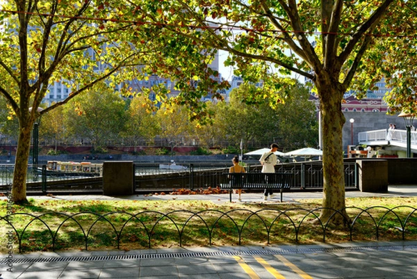 Obraz Shaded trees and a couple sitting on a bench along the Yarra River in Melbourne CBD, Australia, offering a peaceful green space in the city center.