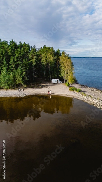 Fototapeta A serene summer day at Lake Saimaa, where a camper van is parked near the shore. Lush green trees frame the peaceful landscape, reflecting beautifully in the calm water.