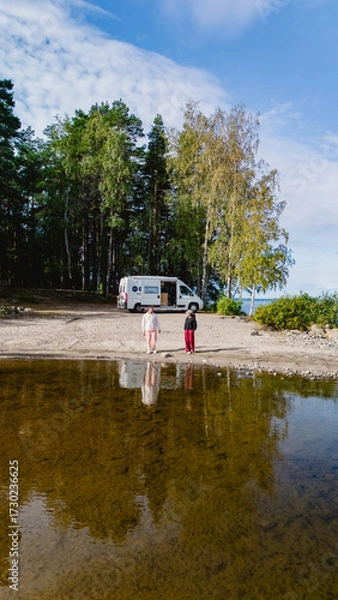 Fototapeta Two individuals stand at the edge of Lake Saimaa, absorbing the serenity of the landscape. A camper van rests nearby, surrounded by vibrant trees under a blue sky, creating a picturesque scene.
