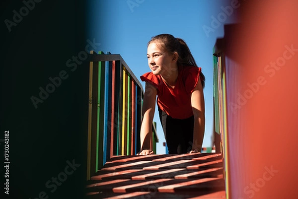 Fototapeta A happy child girl playing on a colorful playground structure in a sunny park, enjoying a fun and active outdoor moment.