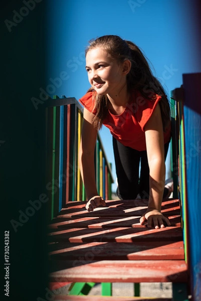 Fototapeta A happy child girl playing on a colorful playground structure in a sunny park, enjoying a fun and active outdoor moment.