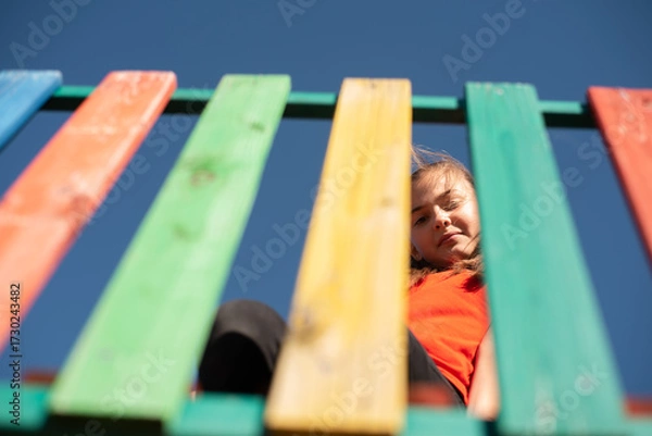 Fototapeta A happy child looking down through colorful wooden playground slats against clear blue sky, playful perspective. Enjoying a fun and active outdoor moment