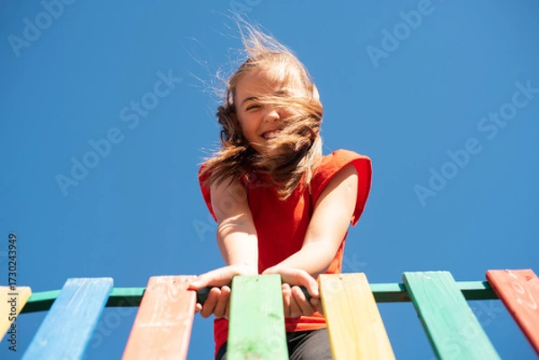 Fototapeta A bright, smiling child girl in a red shirt plays on a colorful wooden playground against a clear blue sky, with her windy hair covering her face, enjoying a fun and active outdoor moment