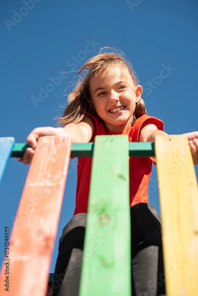 Fototapeta A smiling child in red shirt playing on colorful wooden playground against clear blue sky, enjoying a fun and active outdoor moment.