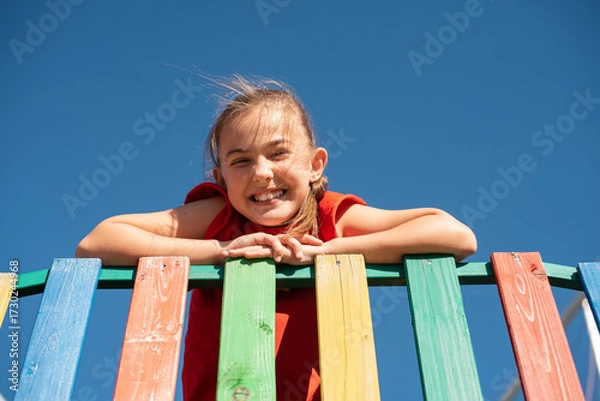 Fototapeta A smiling child in red shirt playing on colorful wooden playground against clear blue sky, enjoying a fun and active outdoor moment.