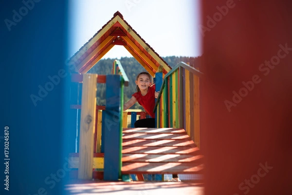 Fototapeta A happy child girl playing on a colorful playground structure in a sunny park, enjoying a fun and active outdoor moment.