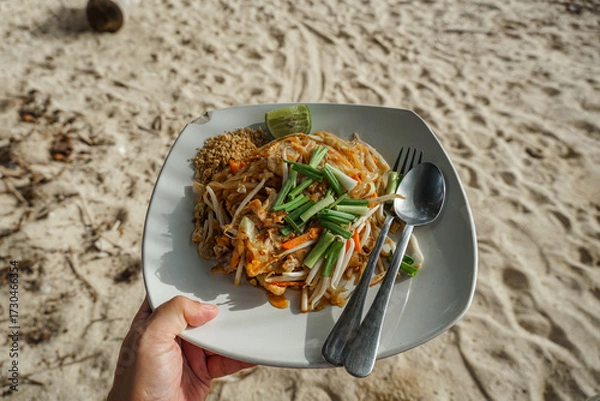 Obraz Top view of a delicious pad Thai dish served at a beachfront on beach sand in the background. A plate of food with pork and lime traditional Thai fried noodle dish.