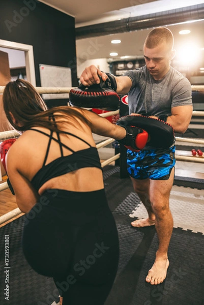 Fototapeta Fitness training session in a boxing gym with a man coaching a woman on technique in the evening