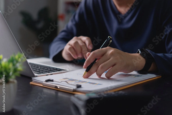 Obraz Businessman analyzing financial data report, hand with pen pointing at chart. Man working with laptop office desk, reviewing business performance, investment growth, and market strategy for success.