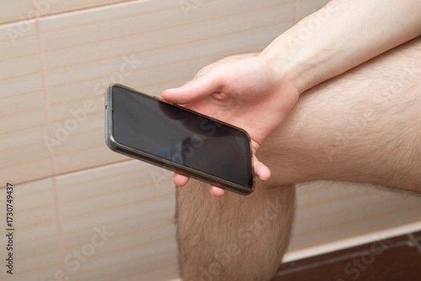 Fototapeta A man is sitting on a toilet with a smartphone.