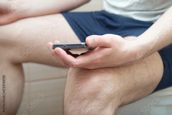 Fototapeta A man is sitting on a toilet with a smartphone.