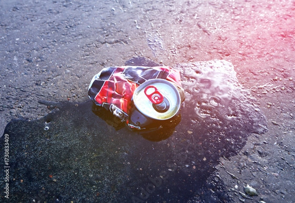 Fototapeta Canned drink spilled on asphalt road on sunset landscape in serene outdoor setting. Beer can lies on wet asphalt. Spilled drink from crumpled Beer Can. Discarded broken drink tin can on asphalt road