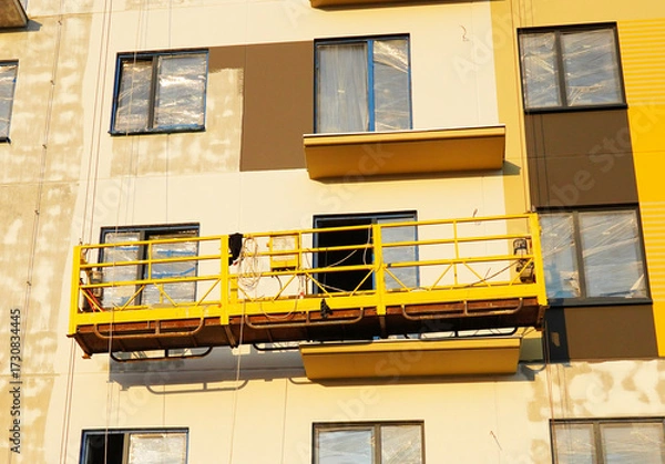 Fototapeta Suspended construction cradle on painting facade of building. Mobile scaffolding platform for construction work at height, facade cleaning, plastering. Insulation and wall painting, cladding.