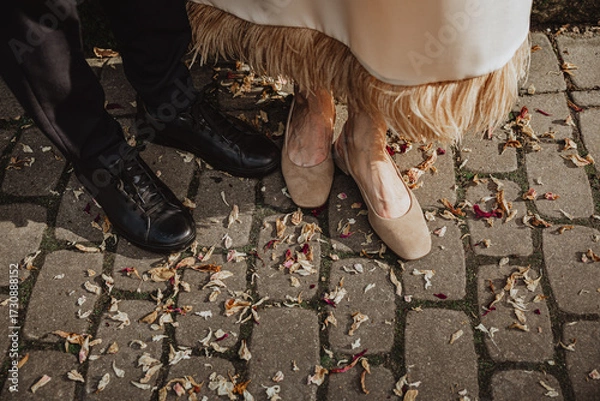 Fototapeta A couple stands close on cobblestone pavement scattered with dried petals, showing contrasting footwear that reflects elegance, unity, and the charm of a special occasion