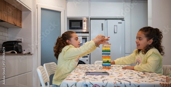 Fototapeta Twin sisters playing a colorful wooden block stacking game together in the kitchen