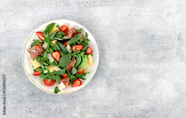 Fototapeta Italian arugula salad with prosciutto, strawberries, melon, cheese on gray background. Top view. Copy space.