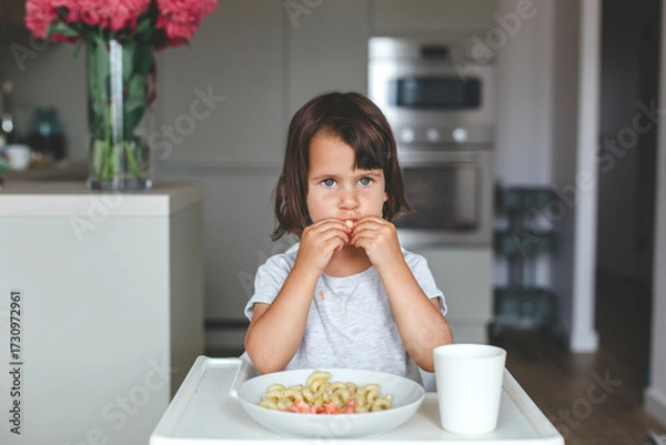 Fototapeta Little girl eating macaroni with both hands, puffing her cheeks while looking ahead, sitting at the table in cozy kitchen, concept of childhood