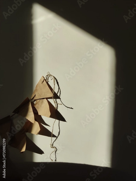 Fototapeta A close-up shot of a rustic, handmade brown paper bunting garland illuminated by a dramatic shaft of late afternoon sunlight against a textured, light-colored wall