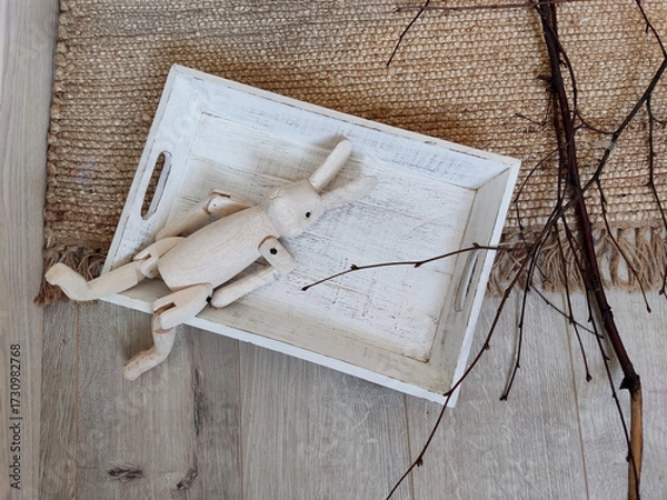 Fototapeta Overhead, flat lay shot of a rustic scene featuring a light-colored wooden rabbit toy with jointed limbs resting inside a distressed white wooden tray, set on a jute rug 