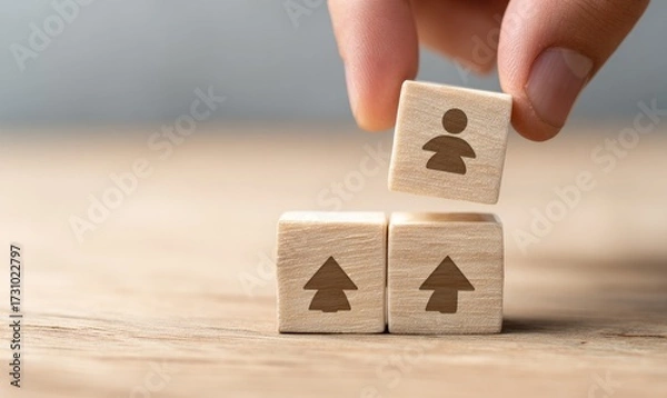 Fototapeta A hand places a small wooden block with a person icon on top of two other blocks with upward-pointing arrows