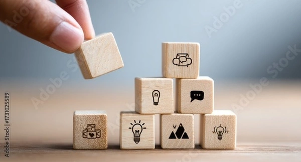 Fototapeta Wooden blocks stacked in a pyramid shape, with icons. A hand is placing a block on top