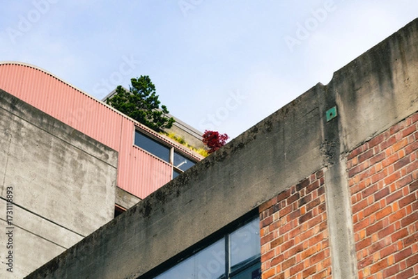 Fototapeta Low angle view of a concrete, red brick, and red-coated metal building exterior, with some balconies that have some plants just barely visible.